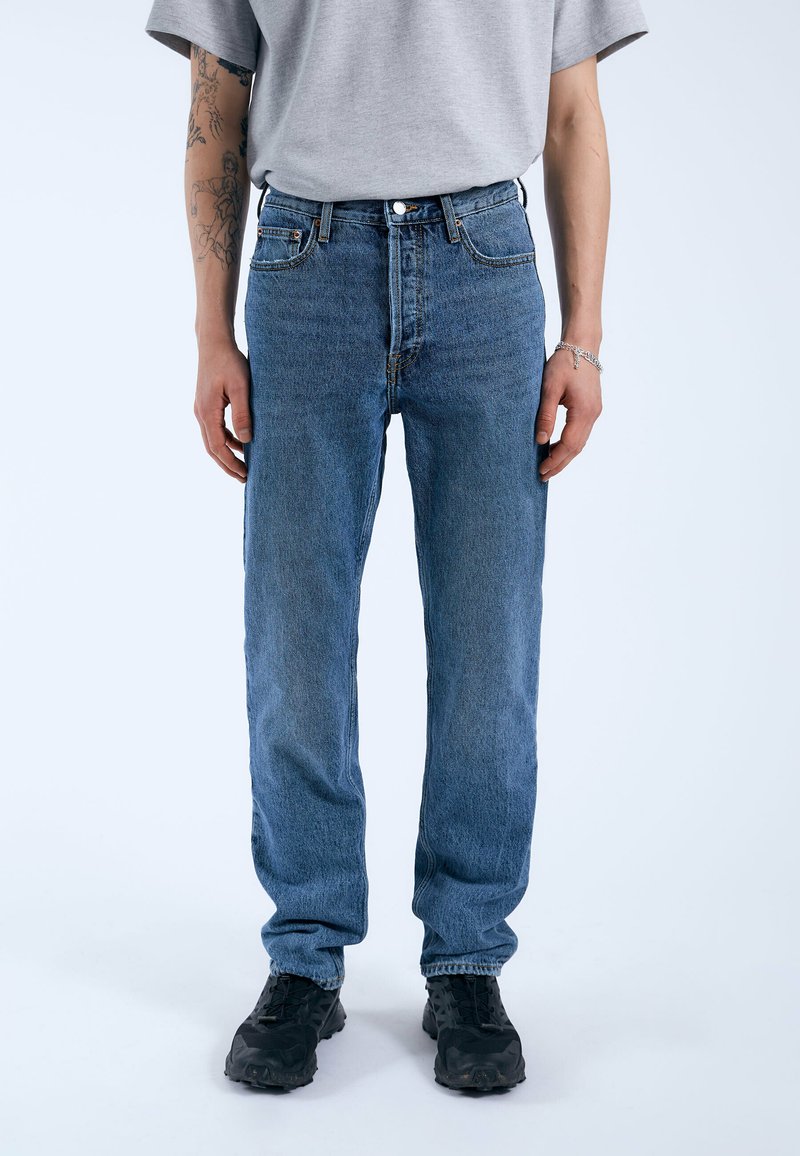 Vaqueros de mezclilla azul con un diseño de pierna recta, que cuentan con cinco bolsillos, un cierre de botón y una textura desgastada. Llevados con zapatillas oscuras.