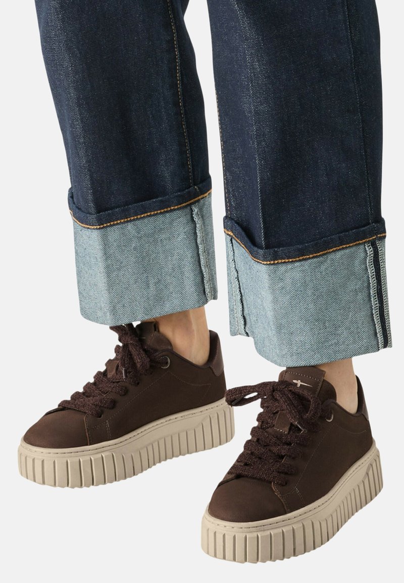 Σκούρα καφέ πλατφόρμα sneakers με χοντρές μπεζ σόλες, φορεμένα με σκούρα τζιν που είναι γυρισμένα στους αστραγάλους και δείχνουν ανοιχτό μπλε εσωτερικό.