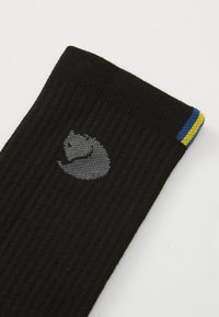 Mustat ribbatussockit, joissa on harmaan ketunpään logo ja pieni sininen ja keltainen raita yläreunassa valkoisella taustalla.
