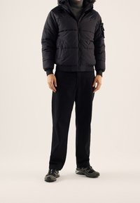 Czarna pikowana kurtka puchowa z kapturem, z przeszywanym wzorem i zamkiem z przodu, w zestawieniu z czarnymi spodniami i szarymi butami sportowymi. Czarne akcenty na kurtce.