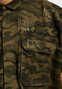 Camicia a motivo camouflage in tessuto verde e marrone, con dettagli sfrangiati, colletto e tasca sul petto con cuciture visibili.