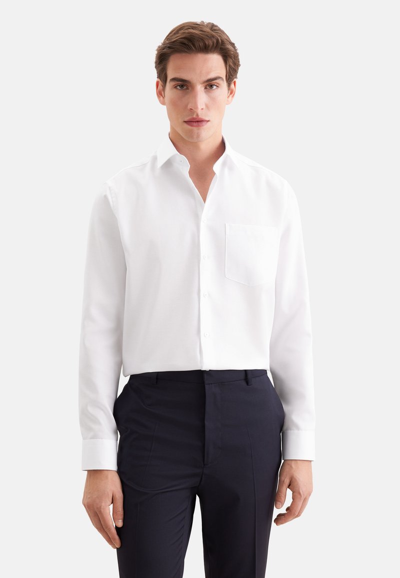 Jeune homme portant une chemise blanche ajustée à manches longues avec une poche poitrine et un pantalon foncé, debout devant un fond uni.