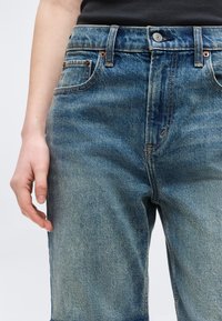 Κοντινό πλάνο ενός ατόμου που φοράει ψηλόμεσο μπλε τζιν με κούμπωμα κουμπιών και φερμουάρ, το αριστερό χέρι άνετα δίπλα στο σώμα.