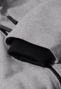 Tissu de veste texturé gris avec une manchette côtelée noire et des détails de fermeture éclair noirs, présentant un motif diagonal subtil.