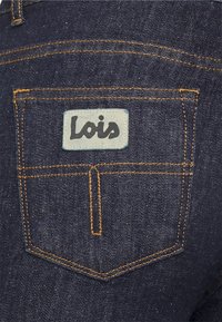 Los jeans de mezclilla azul oscuro presentan un bolsillo trasero con costuras naranjas y una etiqueta rectangular con "Lois" en letras negras.