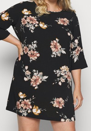 Kobieta ubrana w czarną sukienkę z różowymi, białymi i musztardowymi kwiatowymi wzorami oraz rękawami do łokcia, z ręką opartą na biodrze.