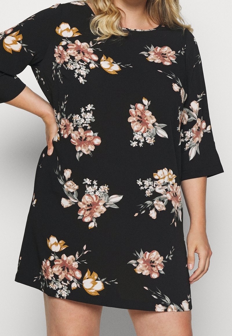 Femme portant une robe noire avec des motifs floraux roses, blancs et moutarde et des manches trois-quarts, la main posée sur la hanche.