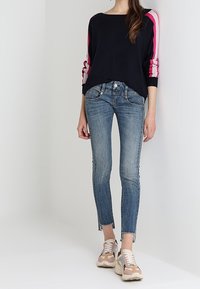 Svart tröja med rosa ränder på ärmarna, kombinerad med ljusblå tighta jeans med fransar i nederkant. Beiga och grå sneakers kompletterar looken.