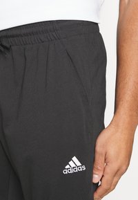 Melni sporta bikses no mīksta auduma ar elastīgu jostasvietu un sānu kabatām. Izcelts balts Adidas logo kreisajā augšstilbā.
