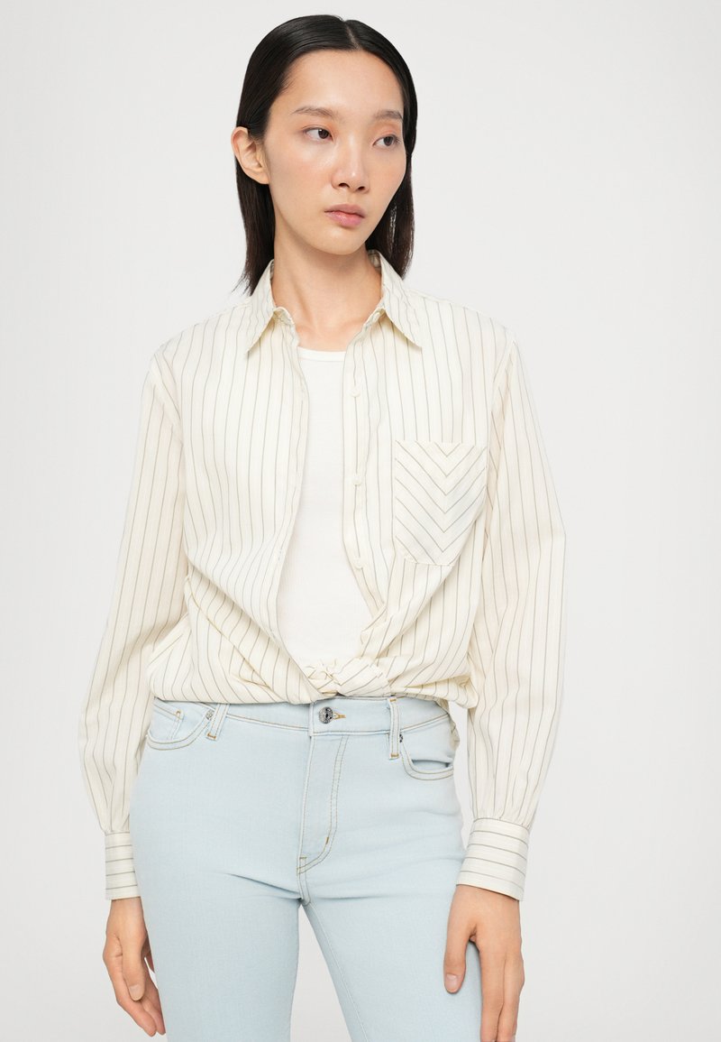 Cremefarbiges Hemd mit dünnen vertikalen Streifen und einer Brusttasche. Kombiniert mit hellblauen, taillierten Jeans. Weicher, leichter Stoff.