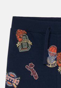 Marina sweatpants med olika färgglada lappar, inklusive en basketboll, en tigerlogotyp och text kopplad till högskolesport.