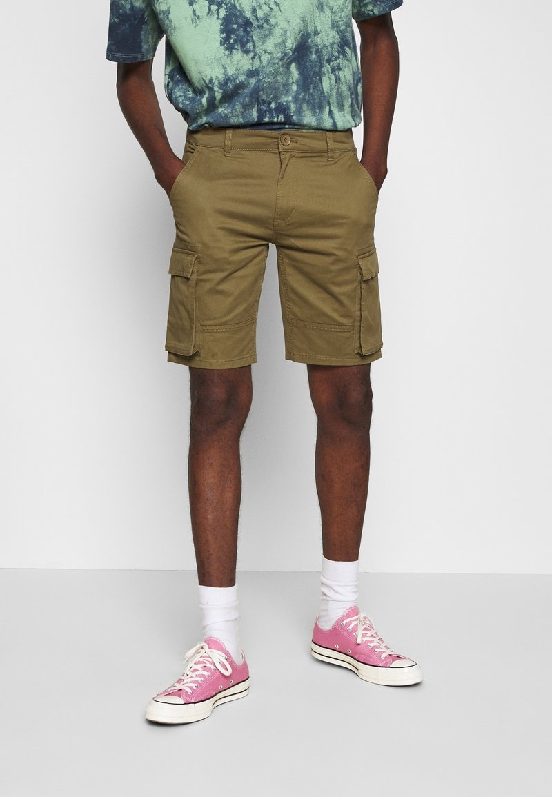 Shorts cargo olive à multiples poches, en tissu mélange de coton, assortis à des baskets roses et des chaussettes blanches, portés avec un t-shirt tie-dye vert et bleu.