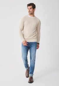 Světle béžový pletený svetr s ribbovaným lemem, kombinovaný s modrými džíny a hnědými koženými kotníkovými botami. Jednoduchý, ležérní outfit.