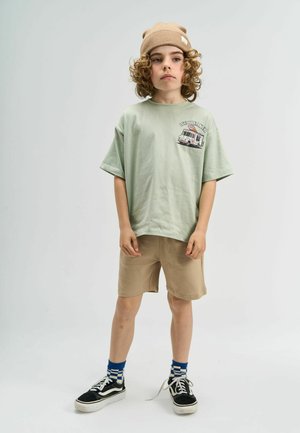 Mündroheline lühikeste varrukatega T-särk, millel on jäätisekäru graafika, millele on sobitatud beežid lühikesed püksid ja mustad tossud siniste ja valgete triibuliste sokkidega.