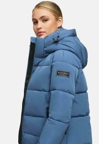 Veste matelassée bleue avec col haut, fermeture éclair et poches latérales. Fabriquée en matériau matelassé avec une finition mate et un patch logo sur la manche.