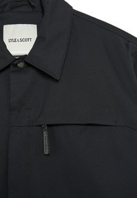 Chaqueta ligera negra con cuello doblado, cierre de snap en la parte delantera y un bolsillo horizontal con cremallera y tirador de borlas. Etiqueta de Lyle & Scott visible.