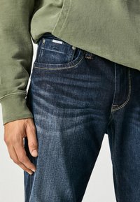 Jeans de color azul oscuro con un ligero desgaste, con costuras en contraste, bolsillo delantero y una etiqueta de marca visible en la cinturilla. Se llevan con un sudadera verde.