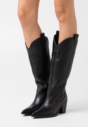 Černé kožené kovbojské boty s špičatými špičkami, širokými podpatky a dekorativním vyšíváním se vzorem ve tvaru vlny. Vysoký svršek s poutky pro snadné nazutí.