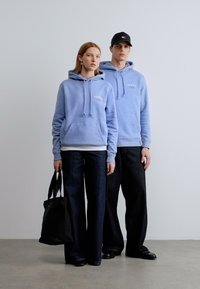 Hellblaue Kapuzenpullover mit Vordertaschen, kombiniert mit dunklen Jeans mit weitem Bein. Eine Person hält eine schwarze Tragetasche.