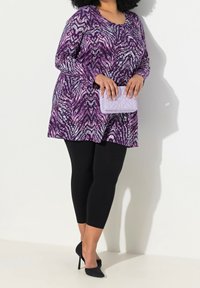 Tunique à manches longues en imprimé animal violet et gris, assortie à des leggings noirs et des talons noirs. Tenant une pochette matelassée violet clair.