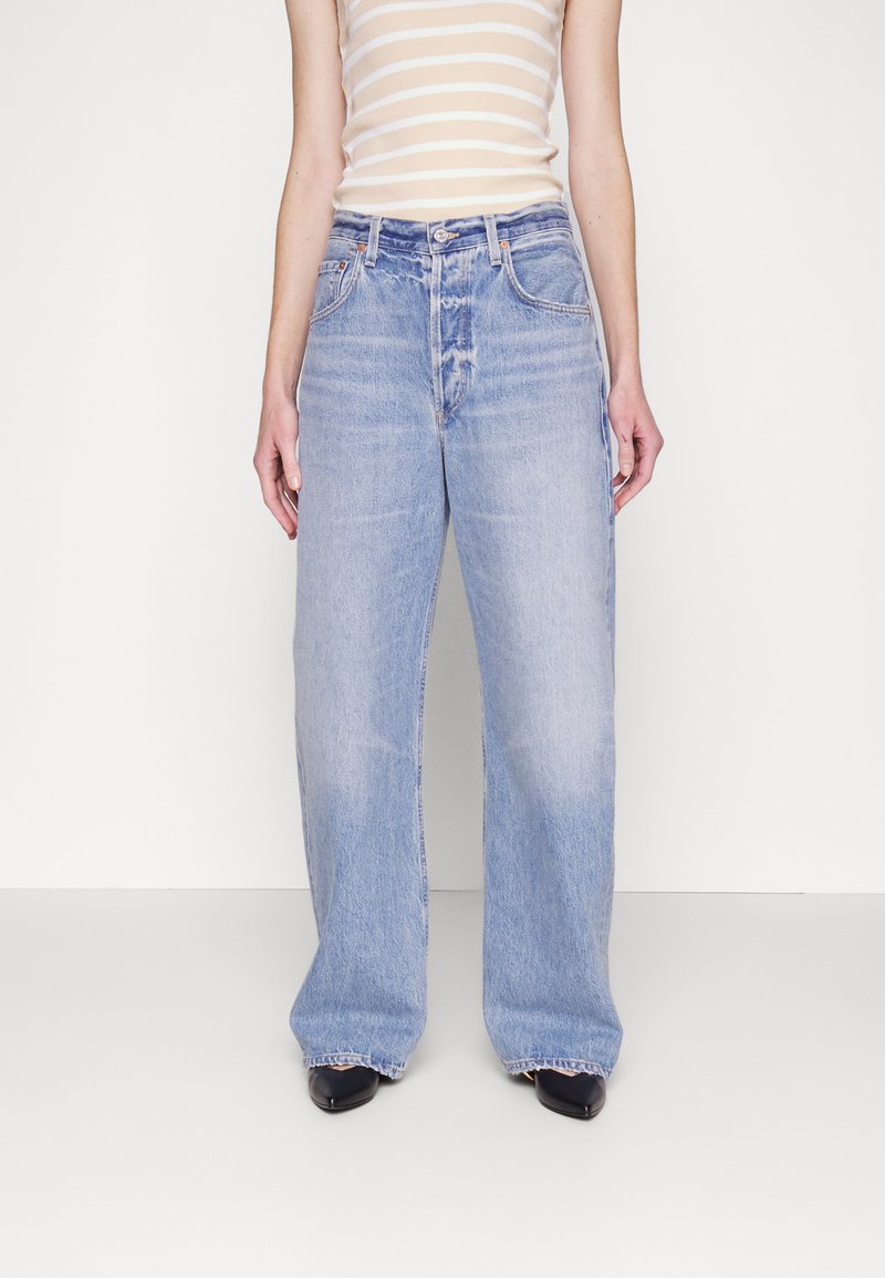 Jean large bleu clair à taille haute, avec un design classique à cinq poches et des ourlets effilochés. Porté avec un débardeur ajusté à rayures.