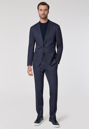 Marineblauw pak met een gestructureerde afwerking, voorzien van een colbert met één knoop en bijpassende broek. Gestyled met een marineblauwe t-shirt en casual sneakers.