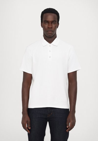 White polo shirt with a classic collar, three buttons, short sleeves, and a straight hem, paired with dark blue denim jeans.