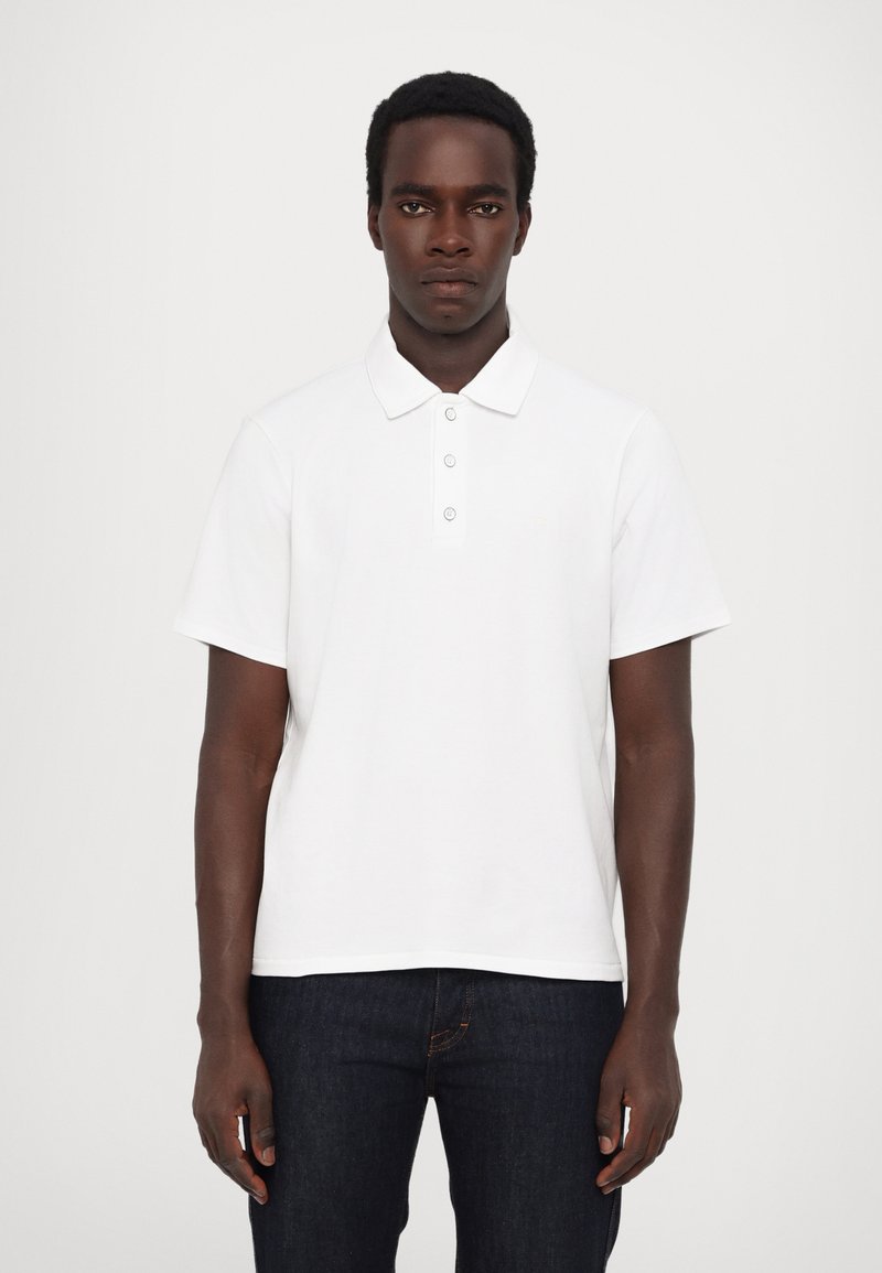 Polo blanc avec un col classique, trois boutons, manches courtes et un ourlet droit, associé à un jean en denim bleu foncé.