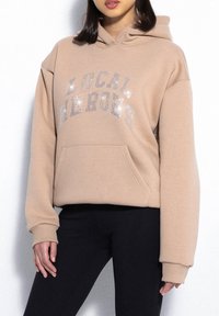 Beżowa bluza z kapturem z napisem "LOKALNE BOHATERY" w srebrnym brokacie. Posiada kieszeń z przodu oraz ściągacze przy rękawach.
