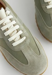 Grüne Wildledersneaker mit einer glatten Textur, cremefarbenen Schnürsenkeln und einer Gummisohle. Sie zeichnen sich durch ein elegantes Design und kontrastierende Materialien aus.