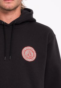 Hoodie noir en coton avec un logo rond rouge représentant une forme de diamant ; comprend des cordons de serrage et des poignets côtelés.