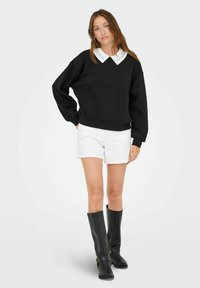 Felpa nera con una camicia bianca a colletto sotto. Abbinata a short bianchi e stivali neri al ginocchio. Design semplice, vestibilità rilassata.