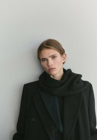 Abrigo negro de lana con diseño cruzado y solapas grandes. Bufanda negra ligeramente texturizada alrededor del cuello. Fondo neutro.