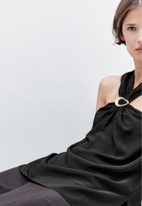 Top nero senza maniche realizzato in tessuto morbido, caratterizzato da un design annodato e un accento circolare dorato sul collo.