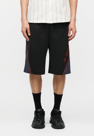 Hombre con pantalones cortos negros y gris oscuro con ribete rojo y escritura roja, calcetines negros, zapatillas negras y una camisa blanca con rayas finas.