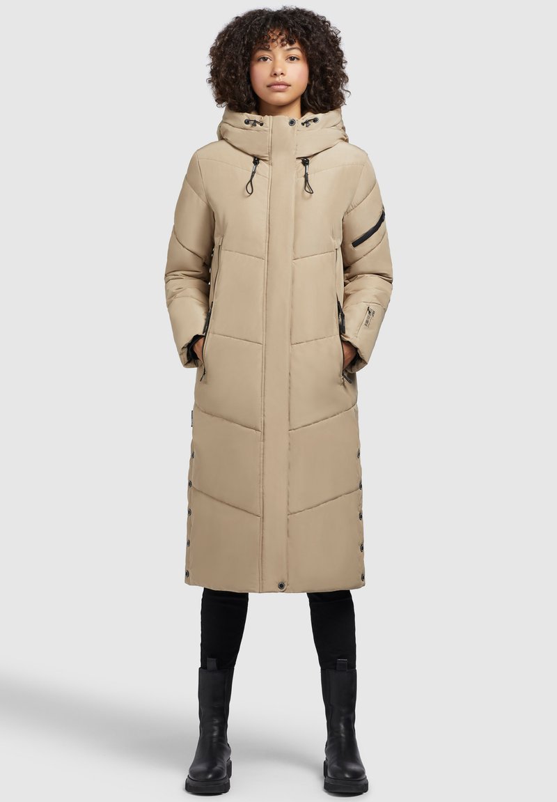 Beige langer Puffermantel mit Kapuze, mit gesteppten Mustern, seitlichen Taschen mit Reißverschlüssen und Druckknopfverschlüssen auf der Vorderseite. Schwarze Akzentdetails.