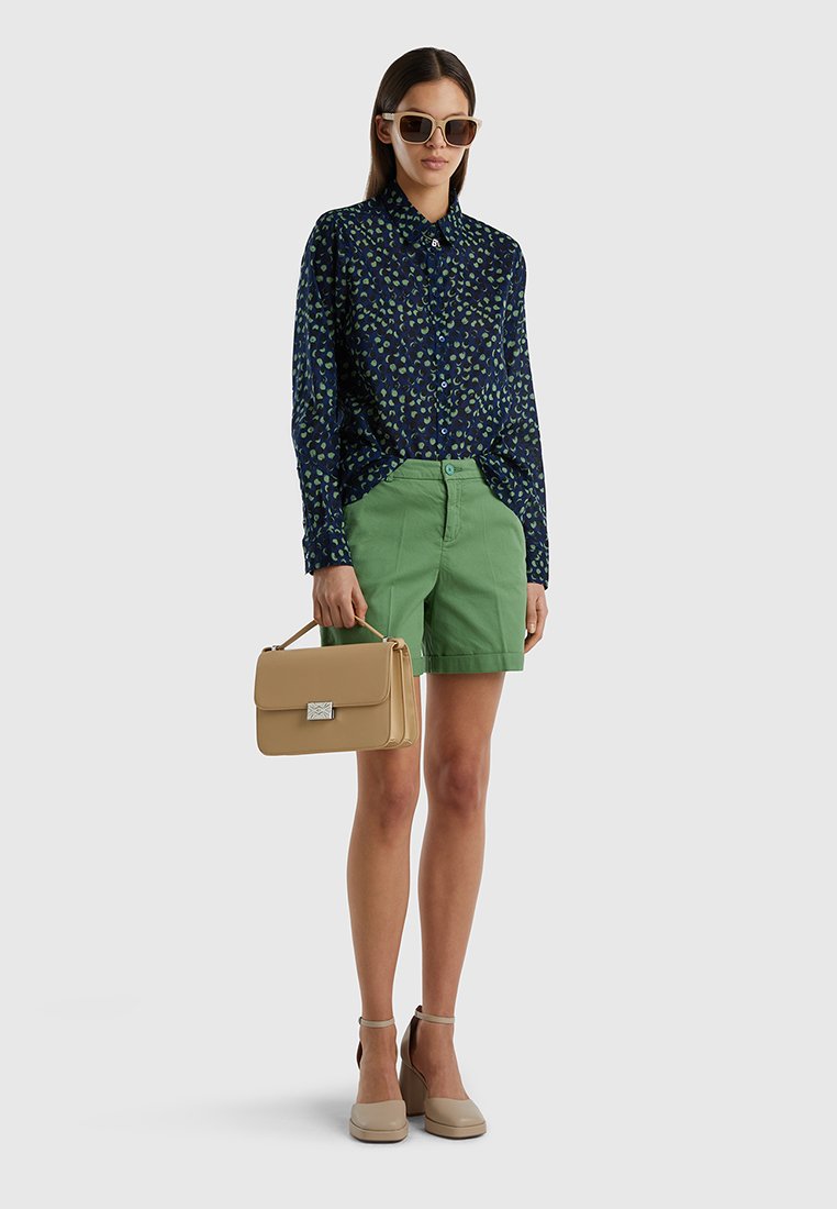 Camicia blu navy con motivo a foglia verde, pantaloni corti verdi, borsa beige con chiusura in argento e scarpe chiare con cinturini alla caviglia.