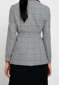 Svart och vit rutig kavaj med en skräddarsydd passform, som har ett bälte i midjan, långa ärmar och en mjuk tygtextur.