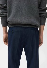 Pulover gri cu dungi, cu mâneci lungi, purtat cu pantaloni în dungi de culoare albastru închis, având buzunare la spate și detalii cu nasturi.