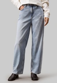 Jeans in denim azzurro chiaro con design a gamba larga, vita alta e dettagli di cucitura visibili. Il modello indossa una maglia bianca e scarpe nere.