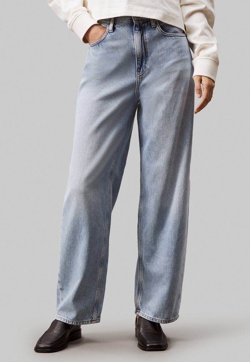 Jeans in denim azzurro chiaro con design a gamba larga, vita alta e dettagli di cucitura visibili. Il modello indossa una maglia bianca e scarpe nere.