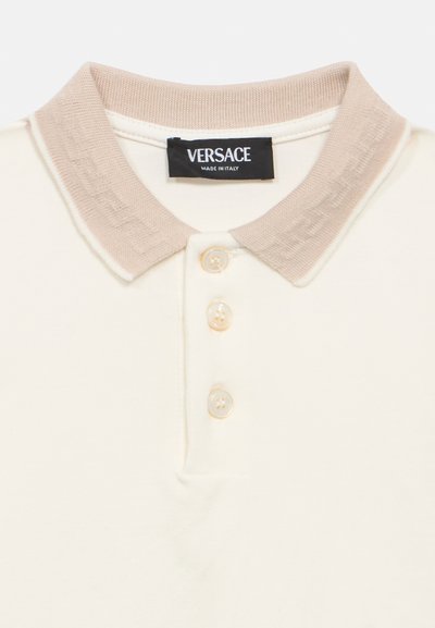 Πουκάμισο πόλο σε κρεμ απόχρωση με απαλό, υφασμάτινο γιακά σε μπεζ χρώμα, που διαθέτει τρία κουμπιά και ετικέτα Versace, τονίζοντας την ποιοτική κατασκευή.