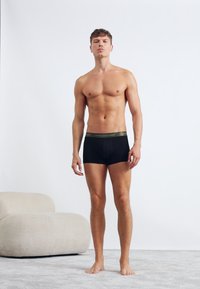 Sorte bomuldsboxershorts med en grøn talje; model står mod en ensfarvet baggrund, hvilket fremhæver pasformen og designet af undertøjet.