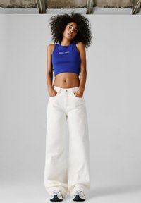 Nainen, jolla on kiharat hiukset, sininen lyhyt toppi ja leveälahkeiset valkoiset housut, seisoo kädet taskuissa yksinkertaisen taustan edessä.