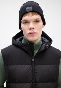 Melna polsterēta veste ar kapuci, ar rāvējslēdzēju priekšā, uzvilkta virs zaļā turtlenecka. Melnā adīta cepure ar reljefu logotipu. Gluda tekstūra, mūsdienīgs dizains.