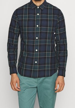Hombre con camisa de cuadros en azul oscuro y verde con botones blancos y pantalones verde claro, de pie frente a un fondo liso.