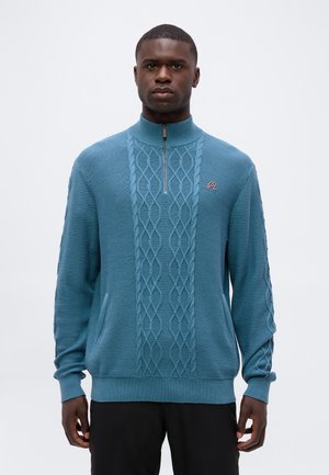 Jeune homme portant un pull bleu torsadé avec un col à fermeture éclair jusqu’au quart et un pantalon noir, debout devant un fond uni clair.