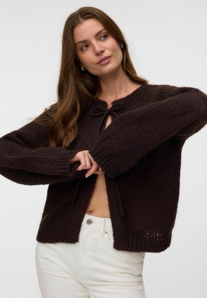 Vrouw met lang bruin haar die een donkerbruine gebreide cardigan draagt, gedeeltelijk gestrikt bij de hals, en een witte broek tegen een effen achtergrond.