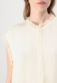 Sleeveless cream blouse with a fringed neckline and smooth texture, featuring a button closure down the front.