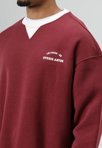 Bordeauxrode sweatshirt met een V-hals, witte kraag en een opvallend "STUDIO ABYSS" logo. Zachte stof met een ontspannen pasvorm en doorgezakte schouders.